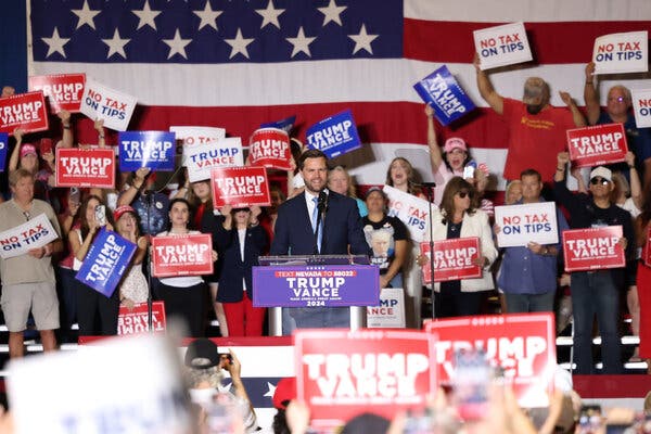 JD Vance stands on a stage speaking at a lectern. Roughly 30 people are on risers behind him holding campaign signs.