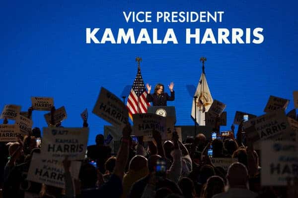 Kamala Harris holds her hands up at a lectern in front of a blue screen. Supporters are waving flags in front of her, and the words “Vice President Kamala Harris” are displayed on the screen behind her.