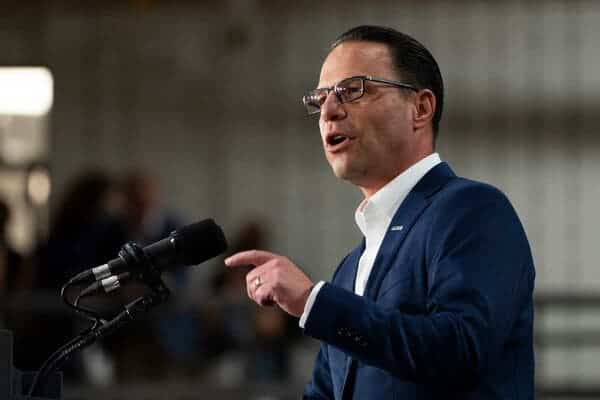 Josh Shapiro speaking in a navy blue jacket and a white shirt with its top button unfastened.