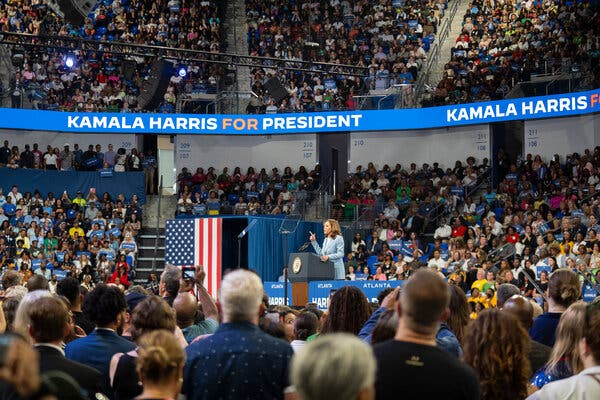 Kamala Harris speaking at a lectern on a stage in the middle of an arena, surrounded by a large crowd. A video board surrounding the stands says Kamala Harris for President.